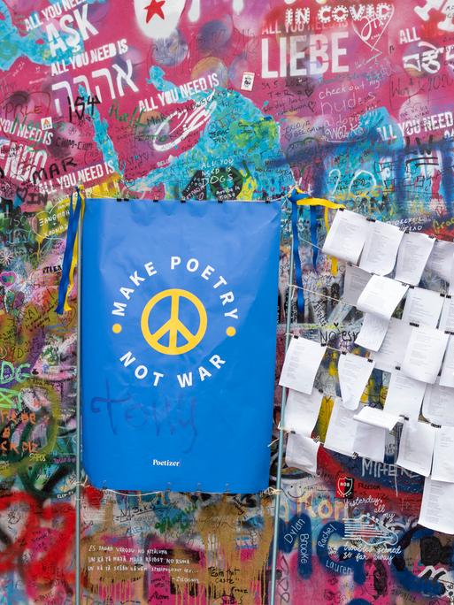 An einer mit Graffiti besprühten Mauer hängen Zettel mit Gedichten. Daneben gelehnt ein blauer Spruchbanner, auf dem in Gelb steht: "Make poetry, not war."