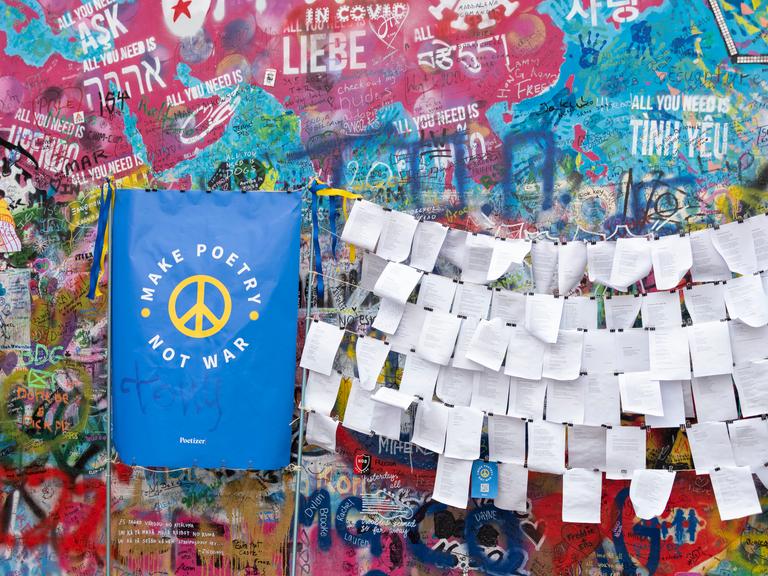 An einer mit Graffiti besprühten Mauer hängen Zettel mit Gedichten. Daneben gelehnt ein blauer Spruchbanner, auf dem in Gelb steht: "Make poetry, not war."