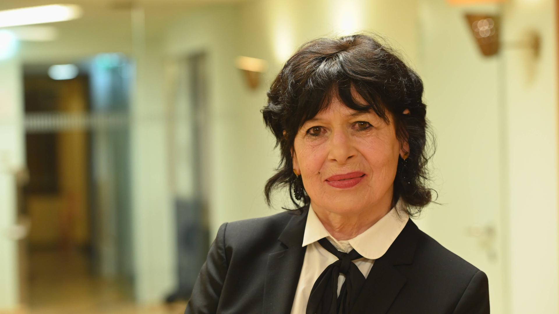 Die Schriftstellerin Monika Helfer schaut leicht lächelnd in die Kamera und trägt einen schwarzen Blazer mit einer weißen Bluse