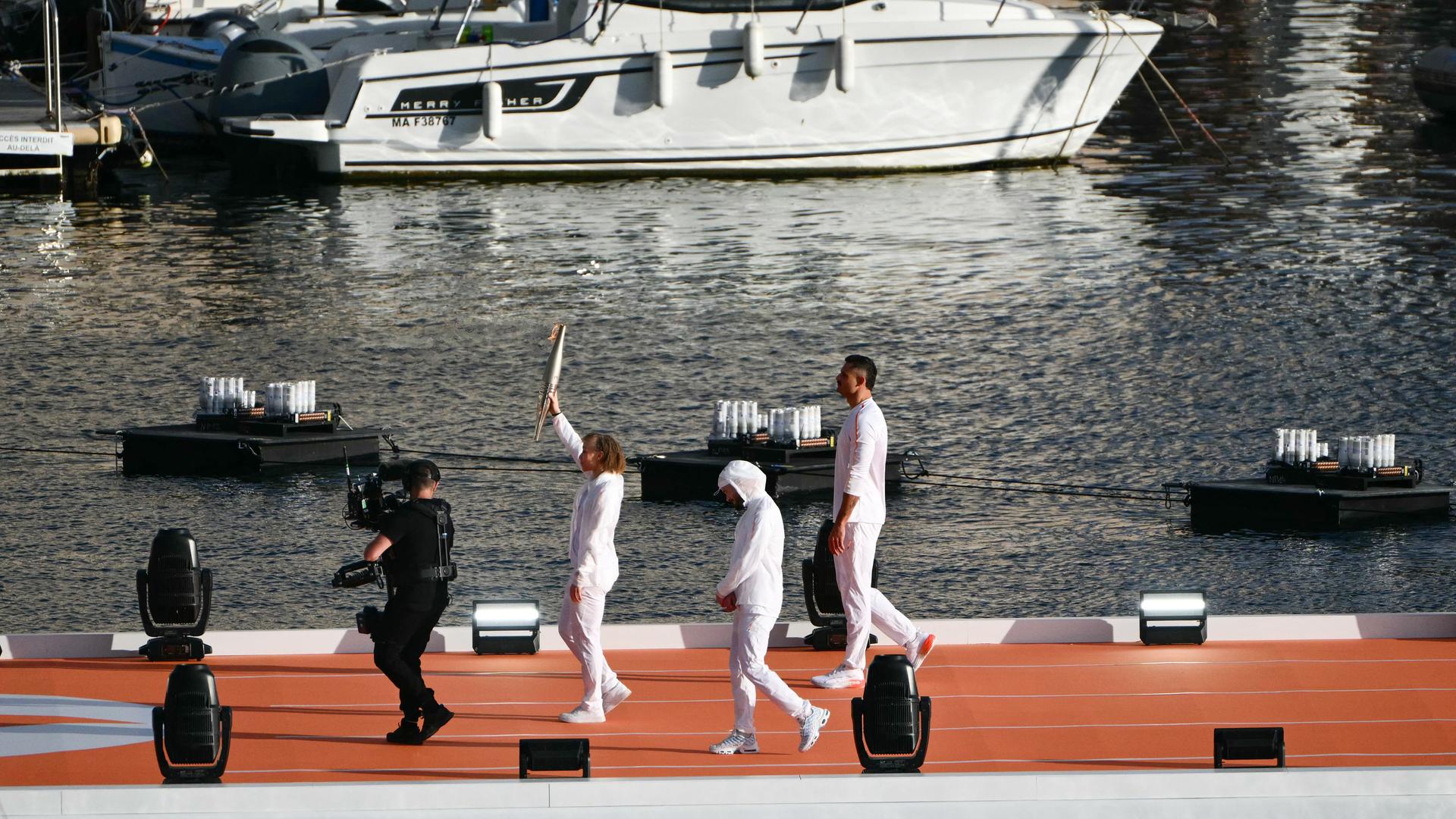 Ein weiß gekleideter Mann trägt die Fackel mit dem olympischen Feuer durch den Hafen. 