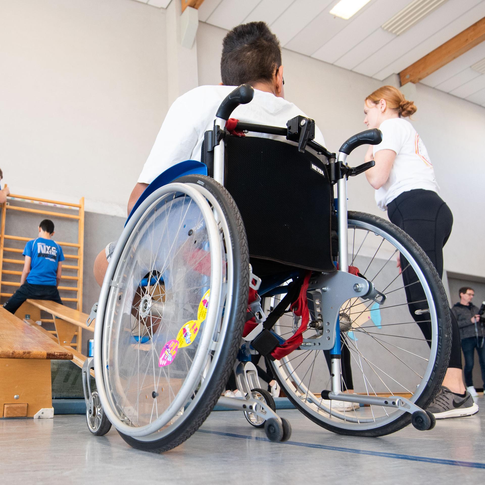 Ein Junge im Rollstuhl und Kinder ohne Behinderung beim Sportunterricht in einer Turnhalle.
