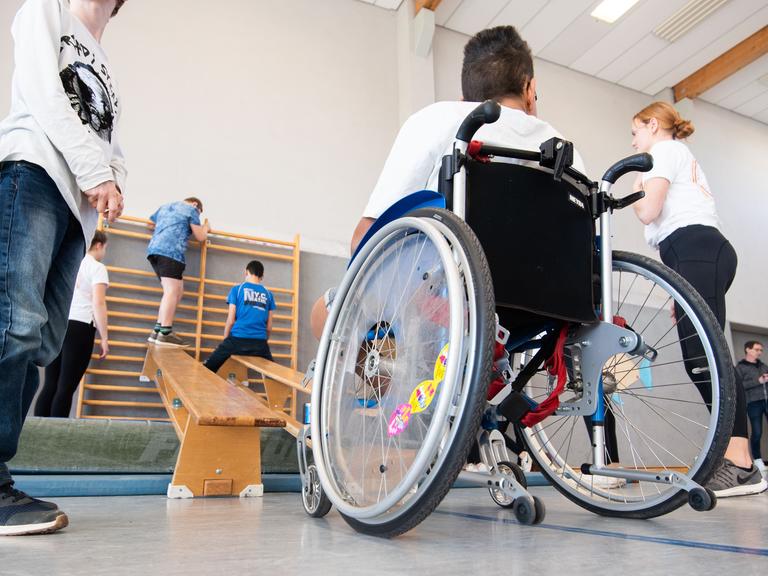 Ein Junge im Rollstuhl und Kinder ohne Behinderung beim Sportunterricht in einer Turnhalle.
