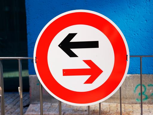Ein Schild weist auf Gegenverkehr und die Vorfahrtsregelung hin. Ein schwarzer Pfeil zeigt nach links, ein in warnendem rot gehaltener Pfeil im rot umrandeten Schild nach rechts.