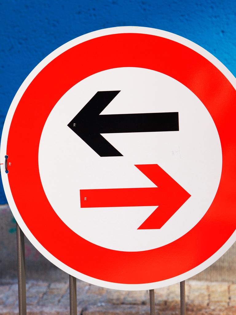 Ein Schild weist auf Gegenverkehr und die Vorfahrtsregelung hin. Ein schwarzer Pfeil zeigt nach links, ein in warnendem rot gehaltener Pfeil im rot umrandeten Schild nach rechts.