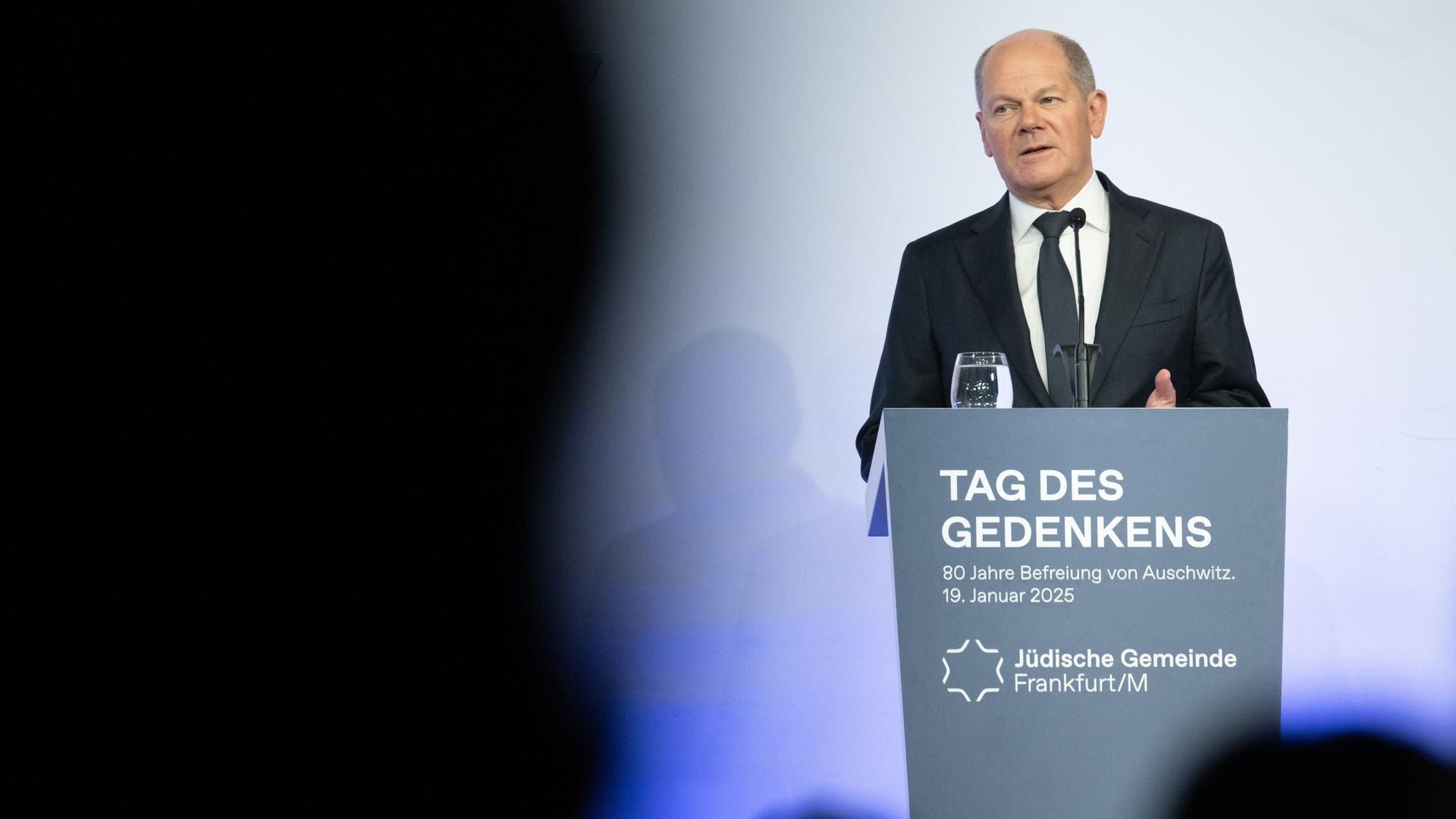 Scholz hält eine Ansprache. Er steht hinter einem Pult und schaut zur Seite. 