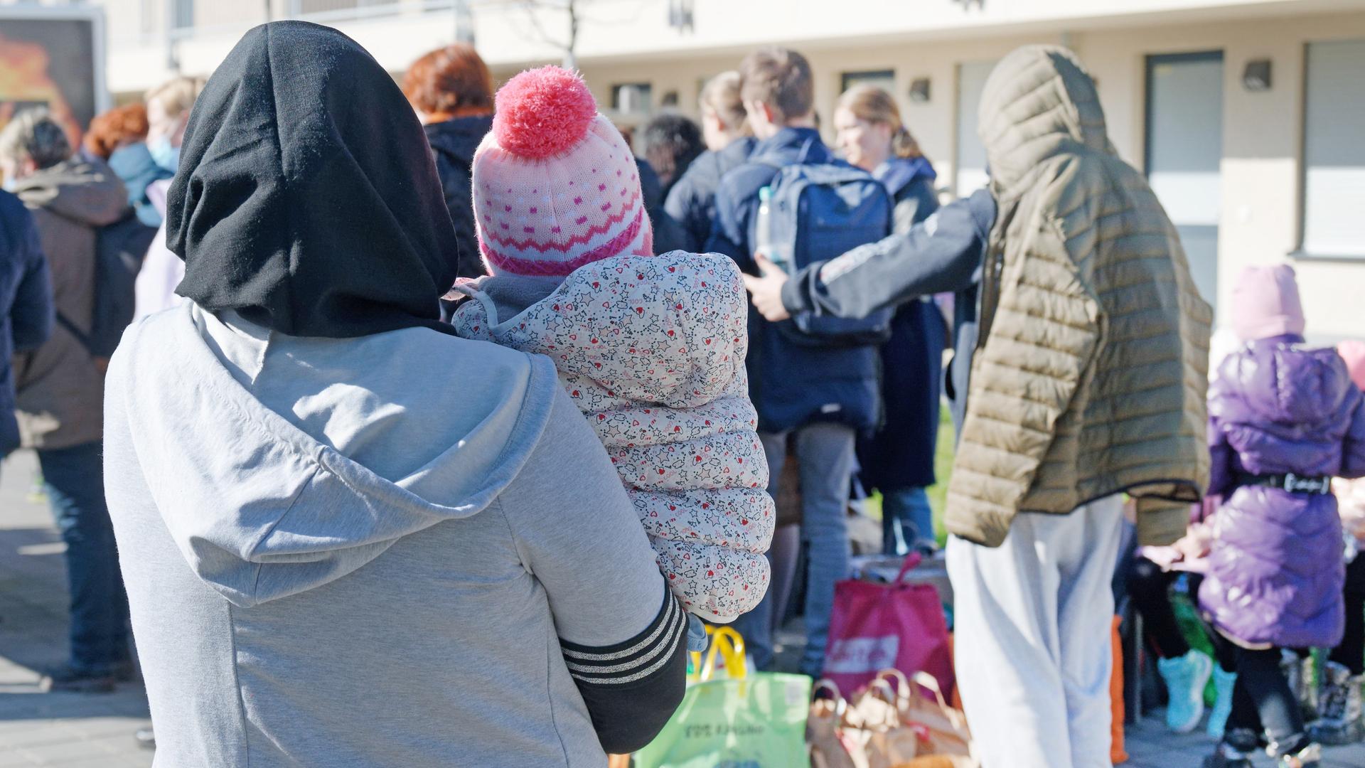 Viele Flüchtlinge sind von hinten vor einem Gebäude fotografiert. Im Vordergrund eine Frau mit einem Kind auf dem Arm.