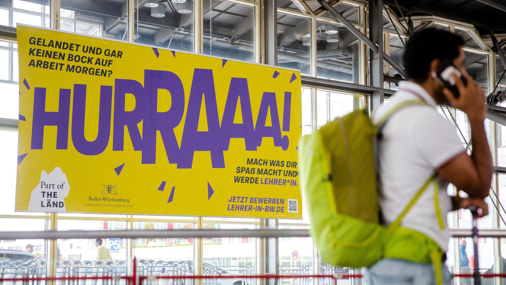 Stuttgart: Ein Werbeplakat für den Beruf des Lehrers hängt am Flughafen in Stuttgart. Davor geht verschwommen ein Mann mit einem Rucksack vorbei, der telefoniert. Stuttgart: Ein Werbeplakat für den Beruf des Lehrers hängt am Flughafen in Stuttgart. Davor geht verschwommen ein Mann mit einem Rucksack vorbei, der telefoniert.