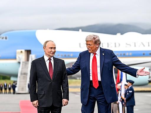Trump empfängt Putin mit einer einladenden Geste seiner Arme vor einem Flugzeug Trump empfängt Putin mit einer einladenden Geste seiner Arme vor einem Flugzeug