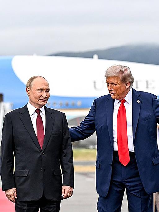 Trump empfängt Putin mit einer einladenden Geste seiner Arme vor einem Flugzeug Trump empfängt Putin mit einer einladenden Geste seiner Arme vor einem Flugzeug