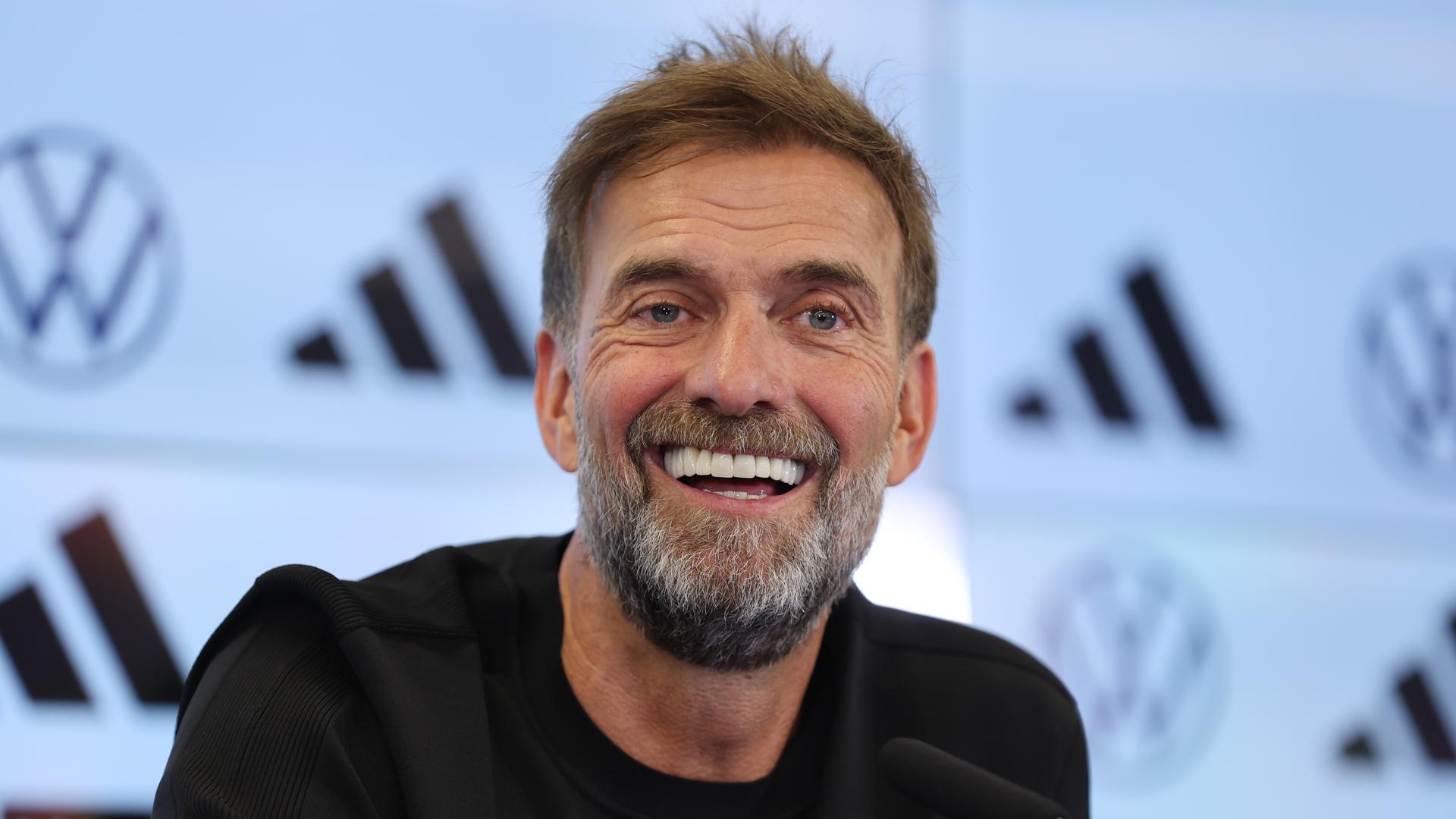 Jürgen Klopp sitzt bei einer Presse-Konferenz. Er schaut an der Kamera vorbei und lacht. An der Wand sind Logos von Marken zu sehen. Jürgen Klopp sitzt bei einer Presse-Konferenz. Er schaut an der Kamera vorbei und lacht. An der Wand sind Logos von Marken zu sehen.