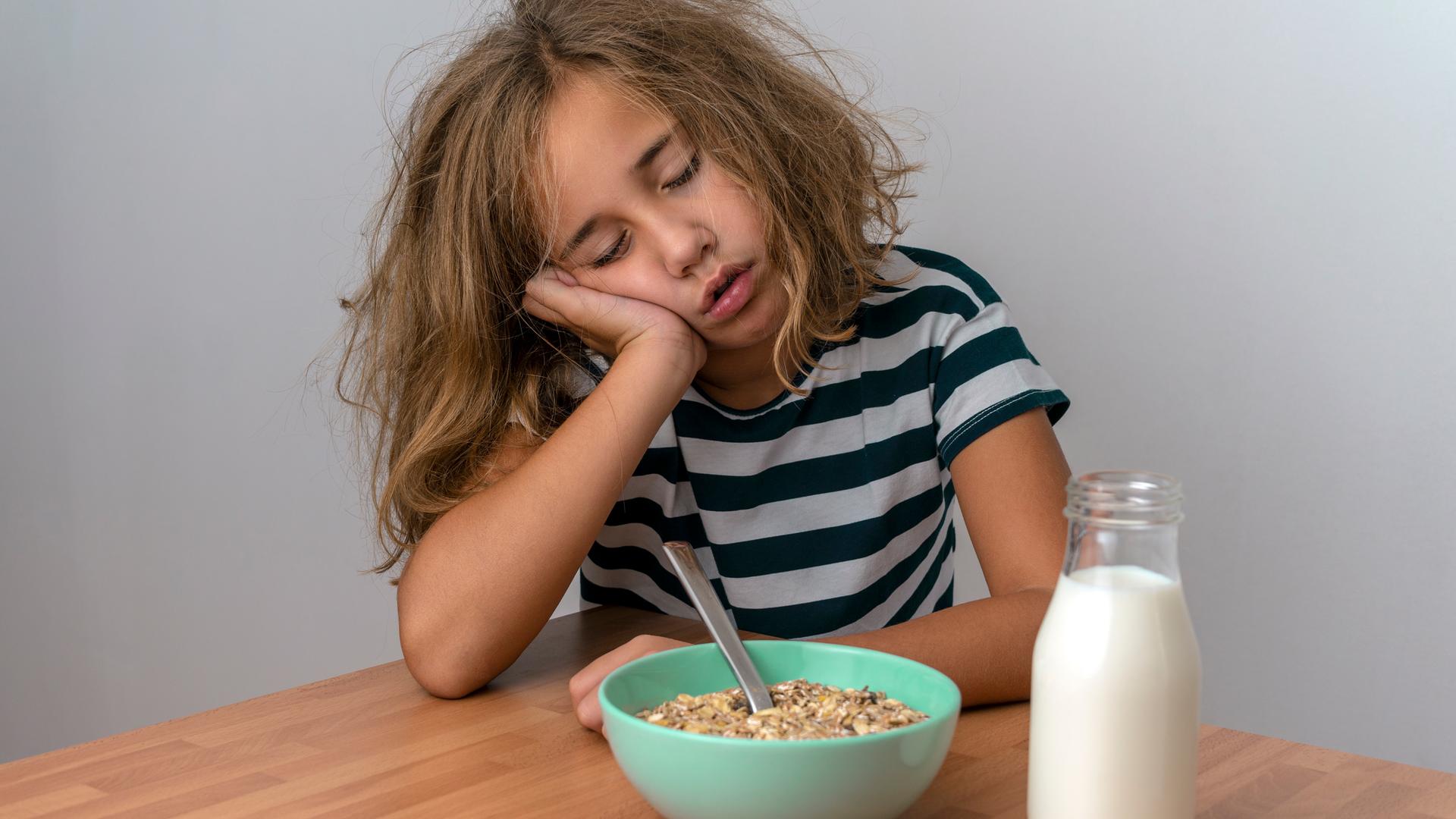 Ein Mädchen sitzt an einem Tisch und schläft über dem Frühstück ein