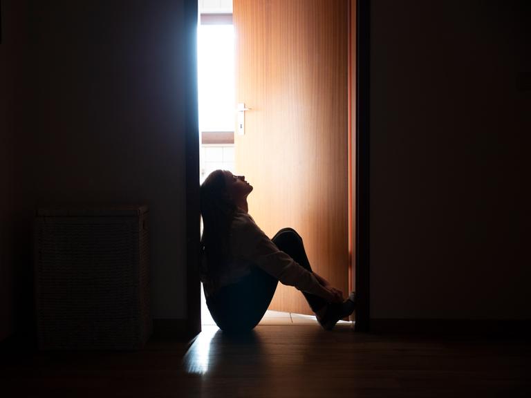 Ein Kind sitzt am Boden in der Dunkelheit. Im Hintergrund öffnet sich eine Tür, durch die ein wenig Licht fällt.