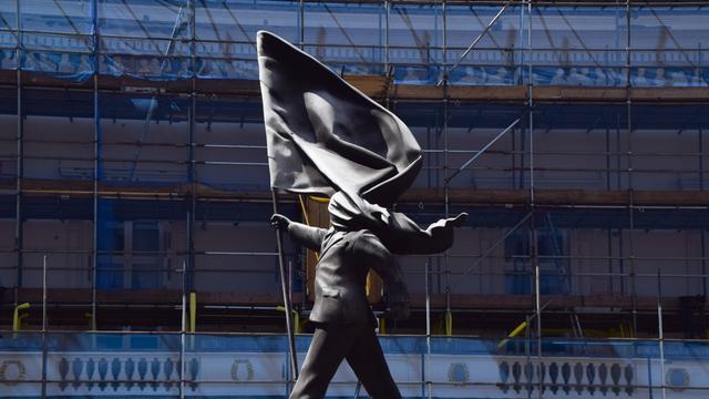 Das Foto zeigt eine Statue in der Stadt London. Es handelt sich um einen Mann, der eine Fahne trägt. Die Fahne verdeckt den Kopf des Mannes. Ein Fuß von dem Mann ist in der Luft.