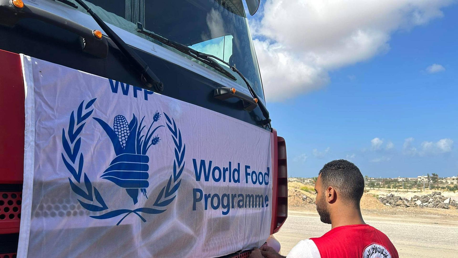 Ein Mann, der eine Weste mit der englischsprachigen und arabischen Aufschrift Ägyptischer Roter Halbmond trägt, befestigt einen Banner an einem LKW mit der Aufschrift World Food Programme.
