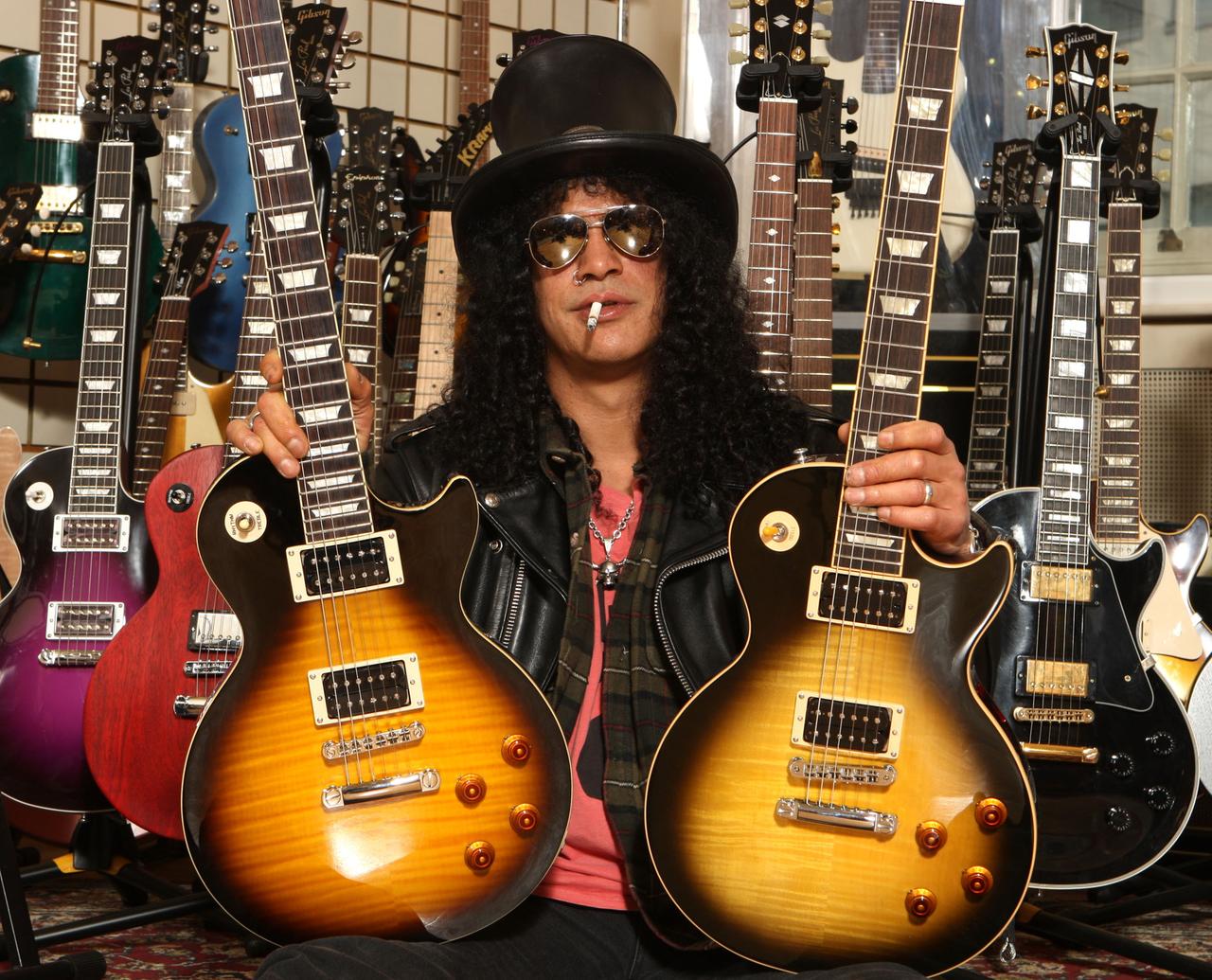 Der amerikanische Musiker Slash posiert mit zwei Gibson Gitarren. Er trägt einen schwarzen Zylinder, eine dunkle Sonnenbrille, hat schwarze lockige Haar und eine Zigarette im Mund.