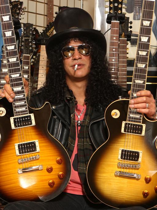 Der amerikanische Musiker Slash posiert mit zwei Gibson Gitarren. Er trägt einen schwarzen Zylinder, eine dunkle Sonnenbrille, hat schwarze lockige Haar und eine Zigarette im Mund.