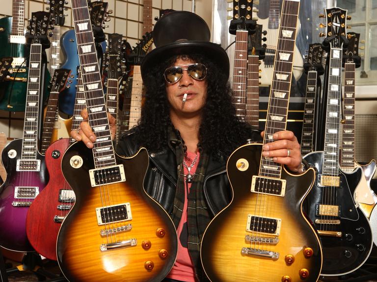 Der amerikanische Musiker Slash posiert mit zwei Gibson Gitarren. Er trägt einen schwarzen Zylinder, eine dunkle Sonnenbrille, hat schwarze lockige Haar und eine Zigarette im Mund.