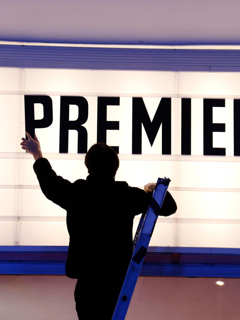 Ein Kinomitarbeiter steht auf einer Leiter und bringt draußen am Kinogebäude den Schriftzug "Premiere" an.