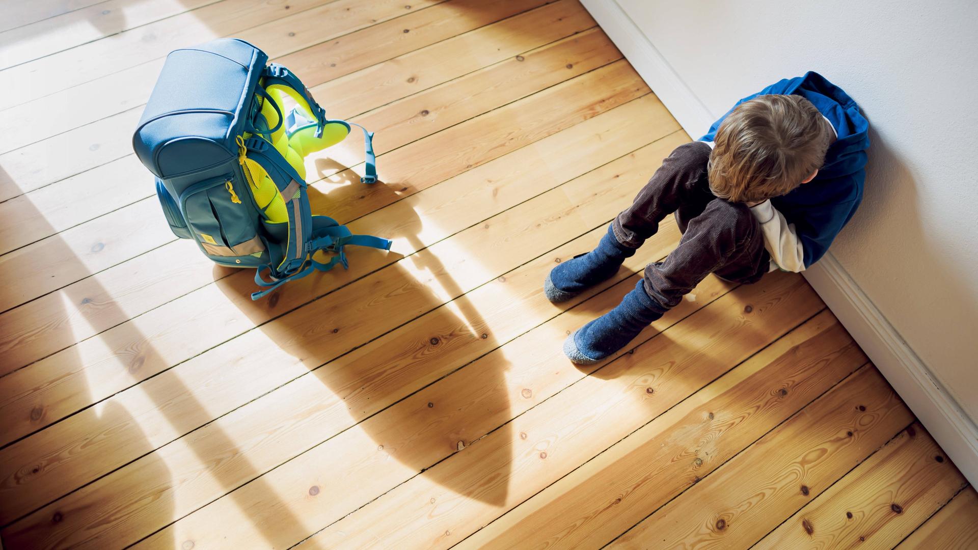 Ein Kind sitzt auf dem hölzernen Fußboden, die Beine angewinkelt, das Gesicht in den Händen vergraben. Neben ihm steht der Schulranzen. Er wirft einen langen Schatten auf den Boden.