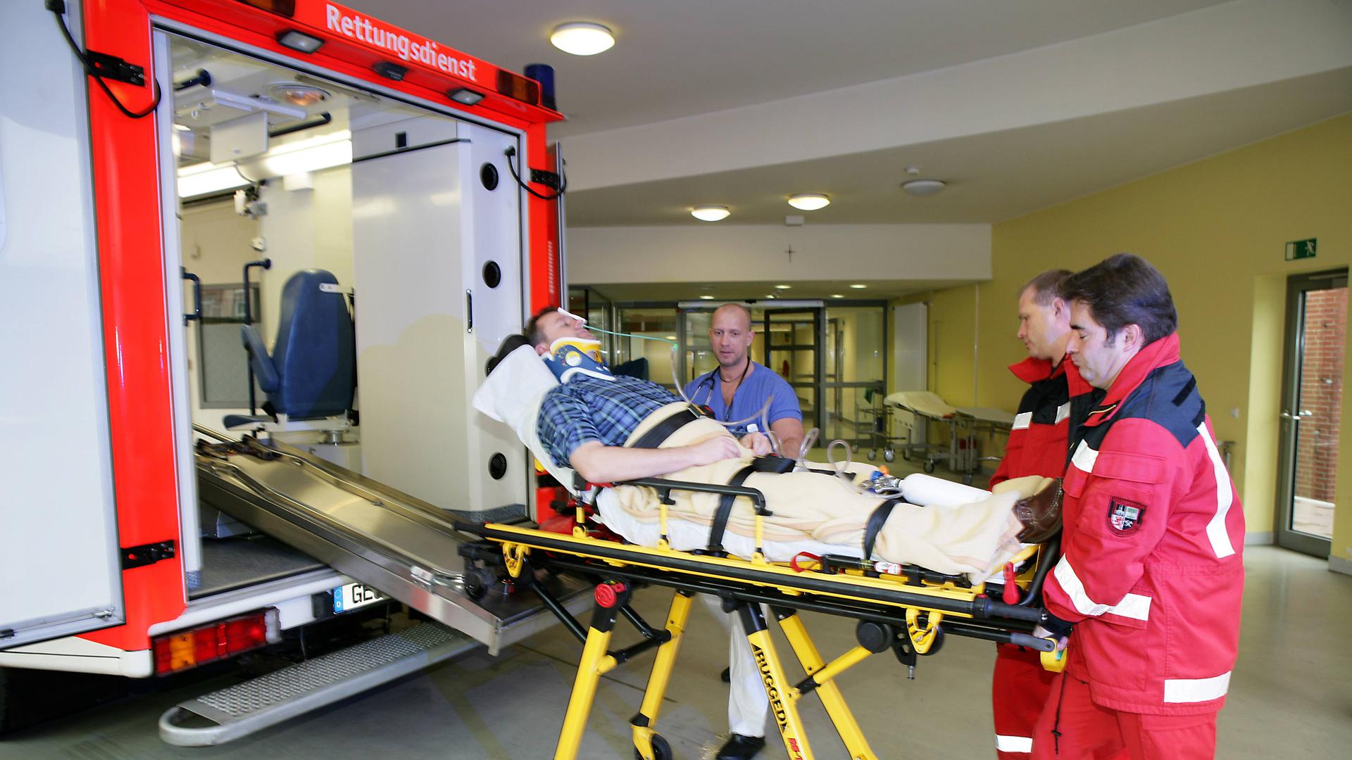 Ein Patient wird aus einem Rettungswagen auf einer Bahre von zwei Männern in roten Overalls in die Notaufnahme eines Krankenhauses gefahren.