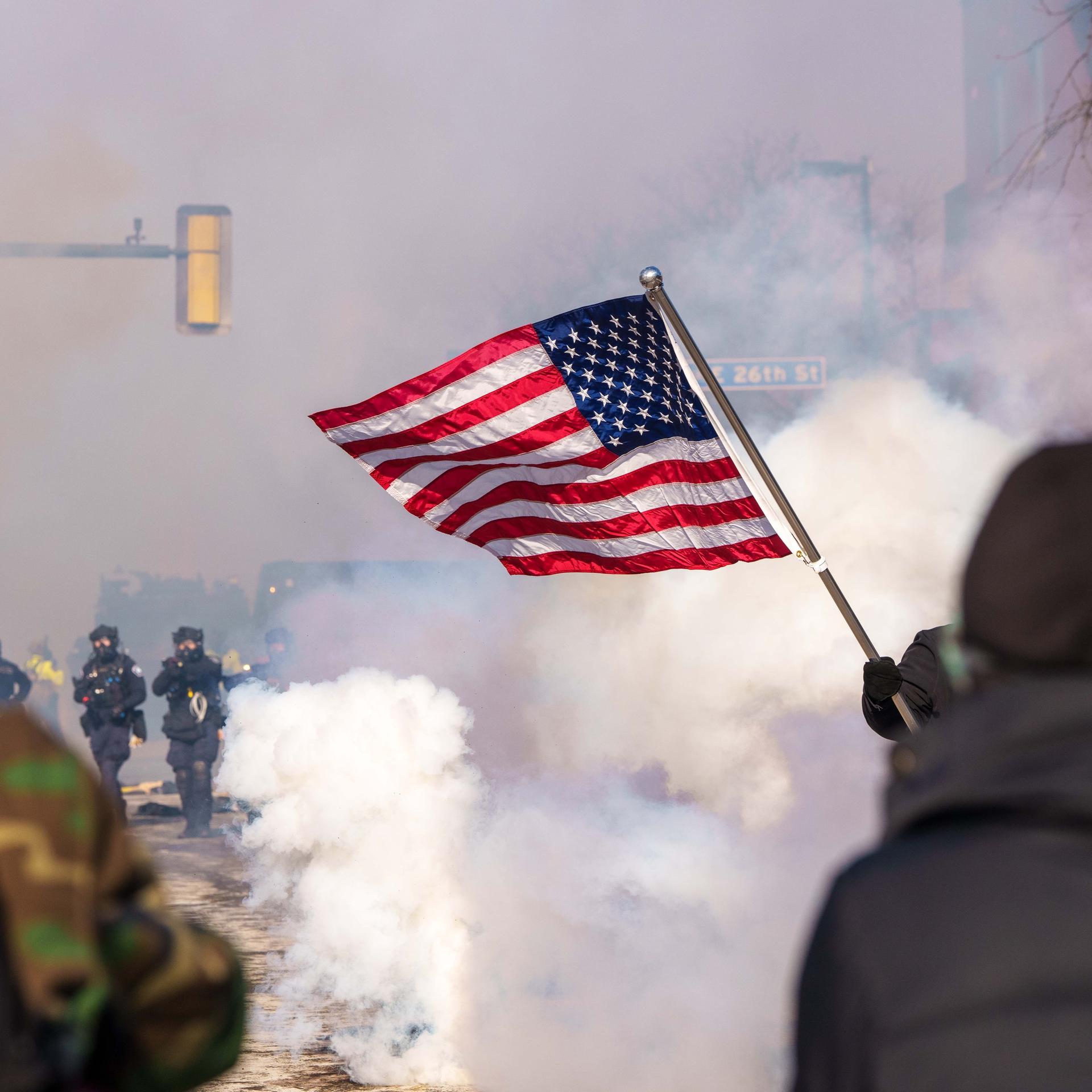 Zwei Protestierende im Vordergrund schwenken eine US-Flagge, während zwei bewaffnete, uniformierte Personen durch Tränengaswolken auf sie zulaufen.