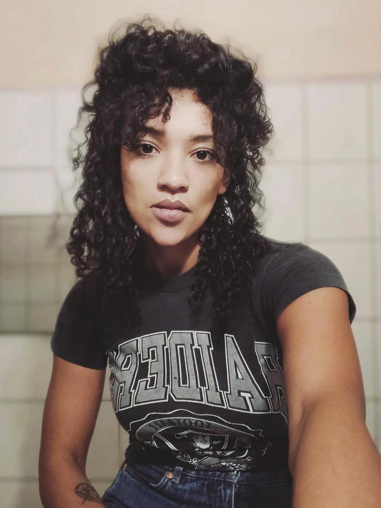 Eine Frau mit langen schwarzen Locken blickt direkt in die Kamera. Sie trägt ein schwarzes T-Shirt und sitzt vor einer gekachelten Wand.