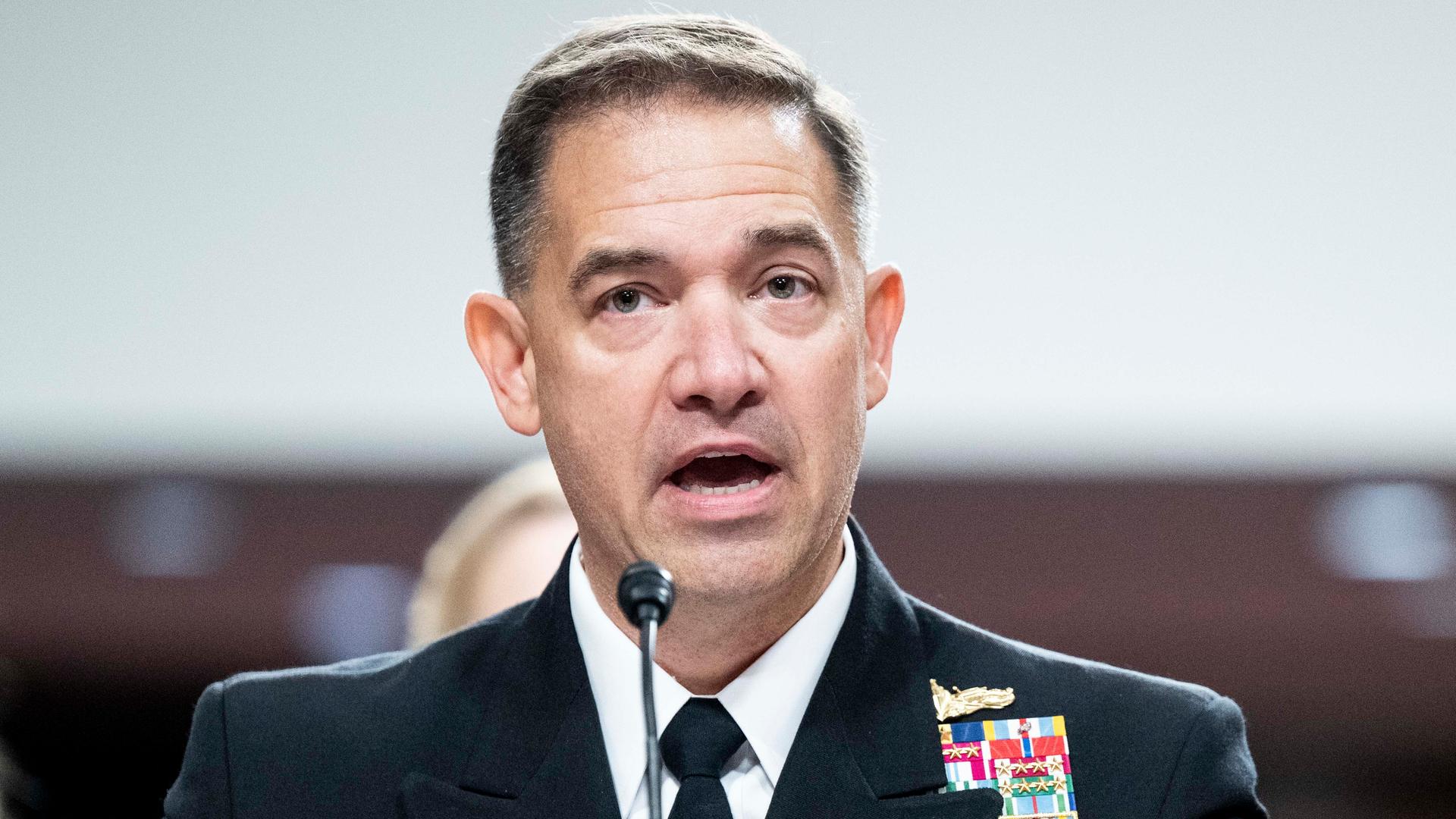 Cooper im dunklen Jackett mit Krawatte spricht gerade in ein Mikrofon. Man sieht ihn von vorne. An seinem Rever haften zahlreiche Auszeichnungen.