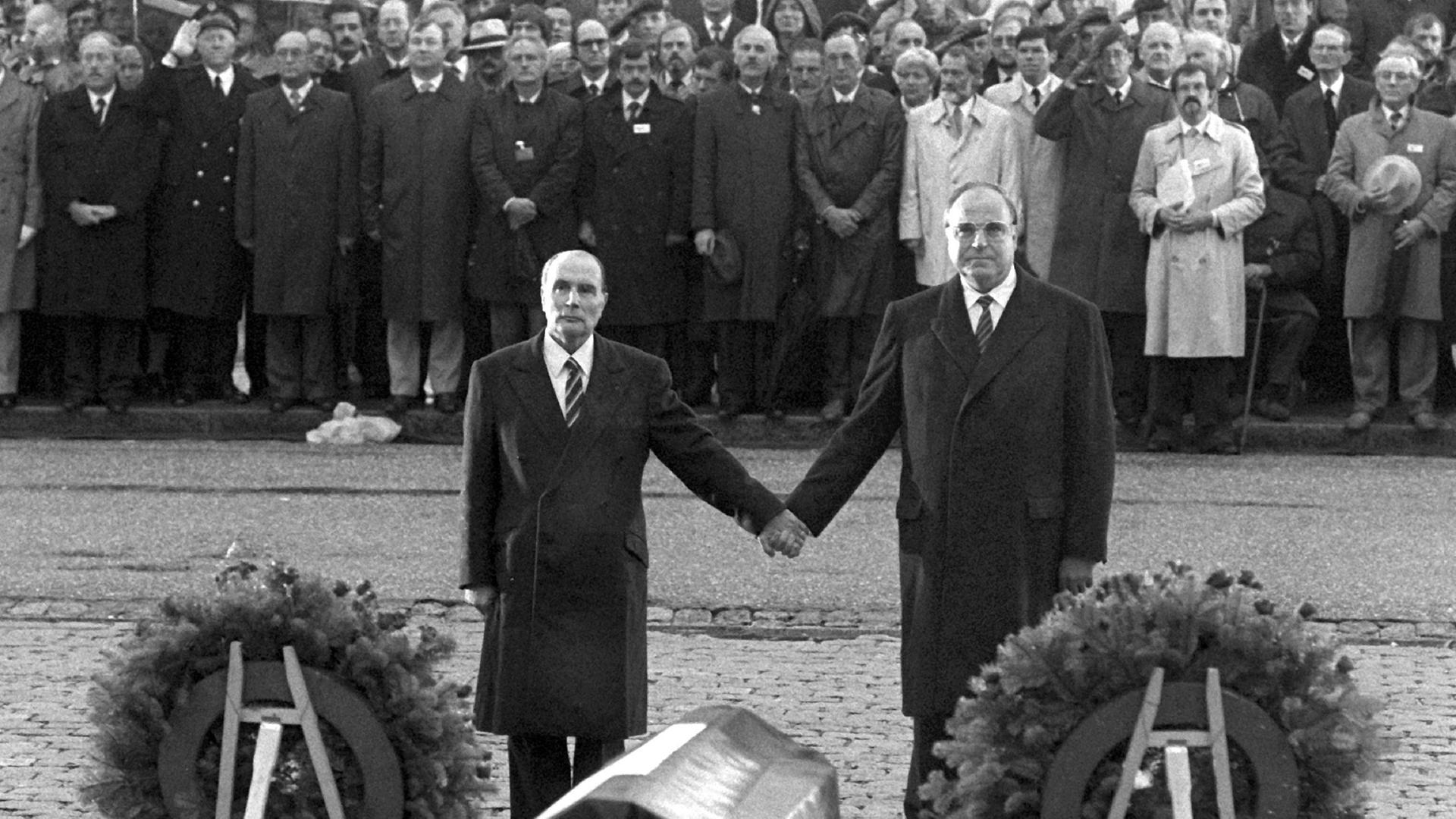 Historisches Schwarzweißfoto von Helmut Kohl und Francois Mitterand, die einander über den Gräbern von Verdun an den Händen halten.