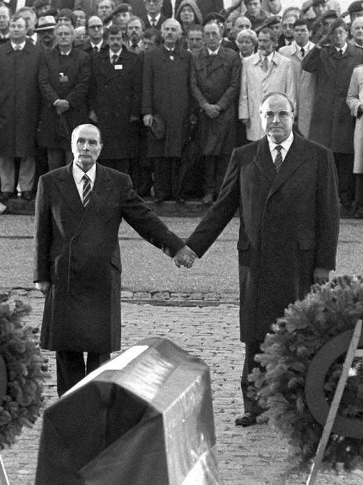 Historisches Schwarzweißfoto von Helmut Kohl und Francois Mitterand, die einander über den Gräbern von Verdun an den Händen halten.