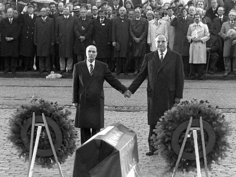 Historisches Schwarzweißfoto von Helmut Kohl und Francois Mitterand, die einander über den Gräbern von Verdun an den Händen halten.