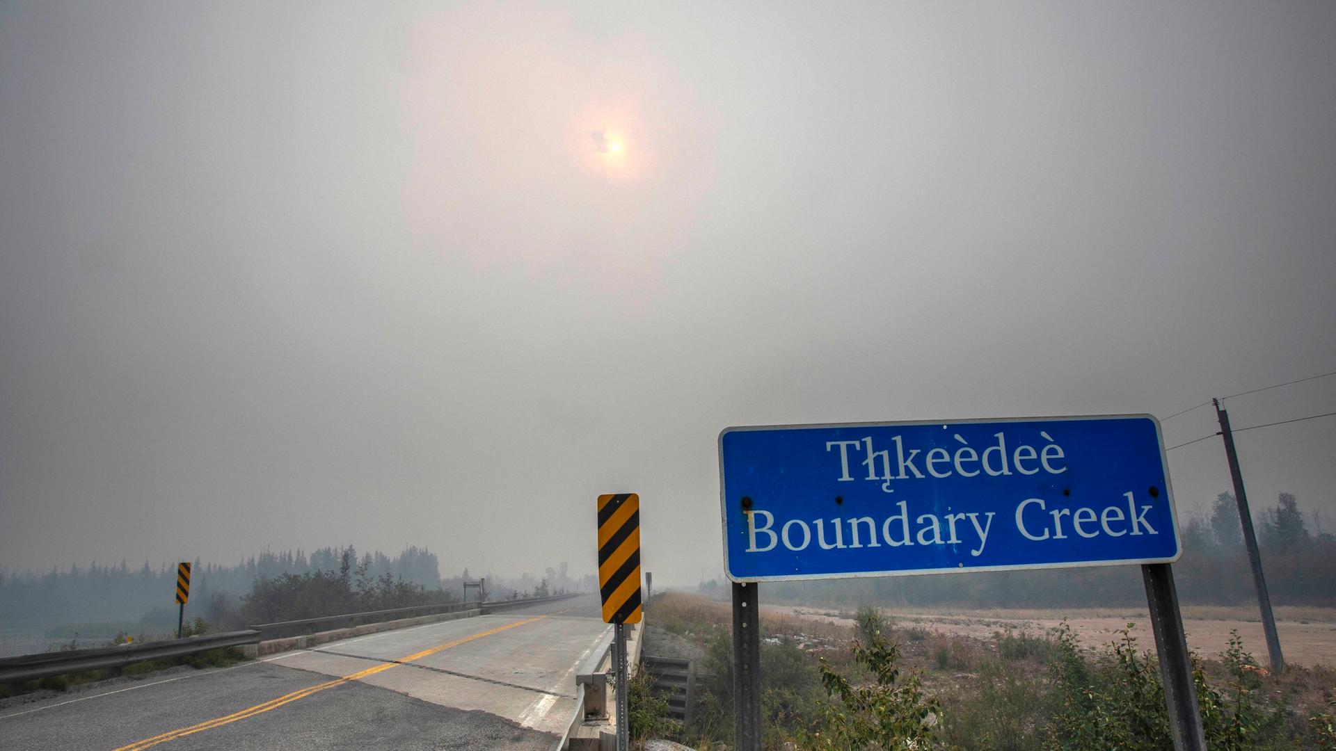 Eine leere Straße, eine vom Rauch der Waldfeuer verhangene Sonne und ein Ortsschild mit dem Namen "Thkeedee Boundary Creek" Eine leere Straße, eine vom Rauch der Waldfeuer verhangene Sonne und ein Ortsschild mit dem Namen "Thkeedee Boundary Creek"