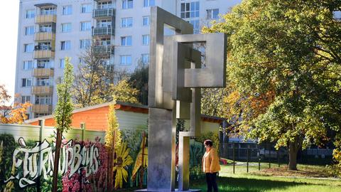 In den Grünanlagen des Wohngebiets Leipzig-Grünau steht eine Frau vor einem kantigen Stahlkunstwerk, im Hintergrund sind Plattenbauten.
