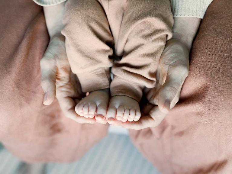 Die Füße eines Babys werden von den Händen einer erwachsenen Person auf dem Schoß gehalten.