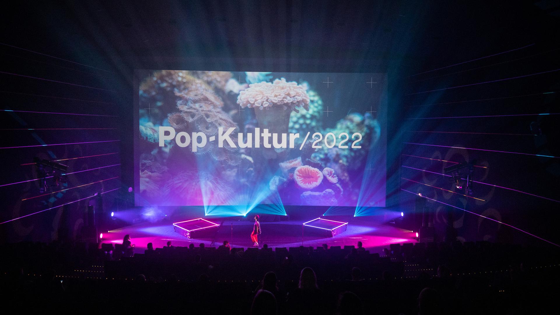 Nuha Ruby Ra steht auf der großen Bühne des Kinos in der Kulturbrauerei, die rot-lila ausgeleuchtet ist und ein Foto einer weißen Koralle im Hintergrund zeigt mit den Schriftzug "Pop-Kultur".