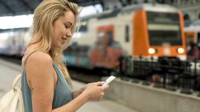 Ein junge Frau wartet am Bahnhof auf einen Zug. Sie sieht dabei auf ihr Handy.