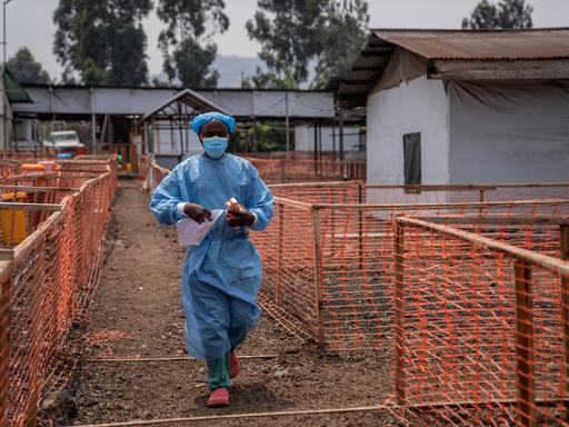 Ein Mitarbeiter in einem Gesundheitszentrum im Kongo. Er trägt einen Mundschutz und einen blauen Plastikkittel. In den Händen hält er Medikamente.