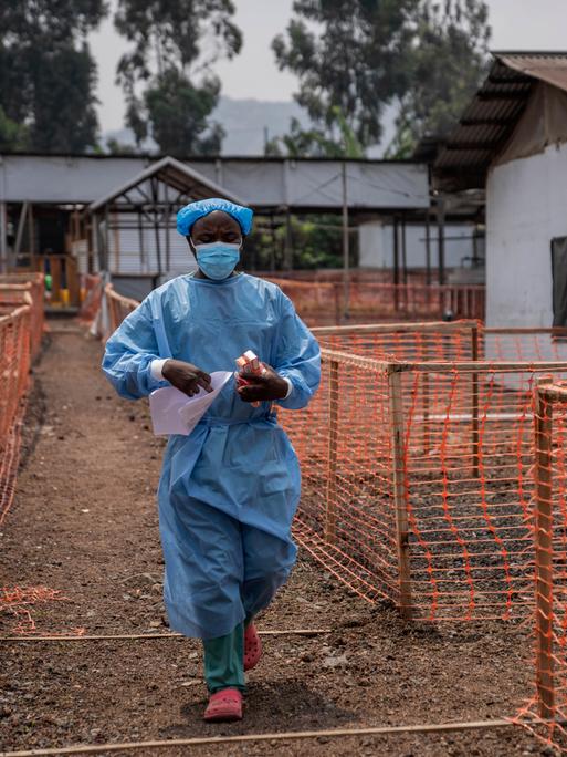 Ein Mitarbeiter in einem Gesundheitszentrum im Kongo. Er trägt einen Mundschutz und einen blauen Plastikkittel. In den Händen hält er Medikamente.