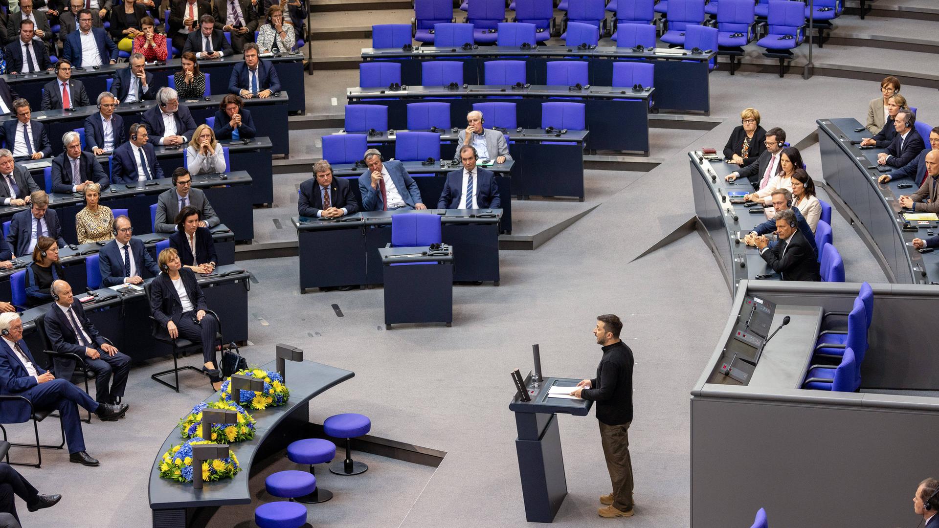 Auf dem Foto sieht man viele leere Plätze im Bundestag. Davor steht Ukraines Präsident Selenskyj am Redner-Pult.