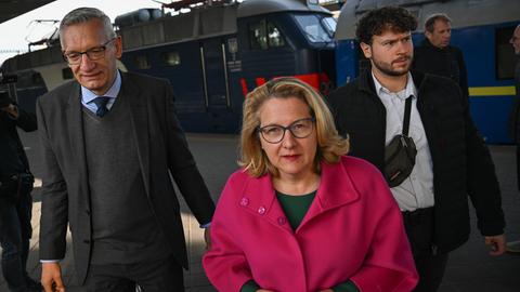 Svenja Schulze steht auf einem Gleis an einem Bahnhof. Hinter ihr stehen zwei Männer.