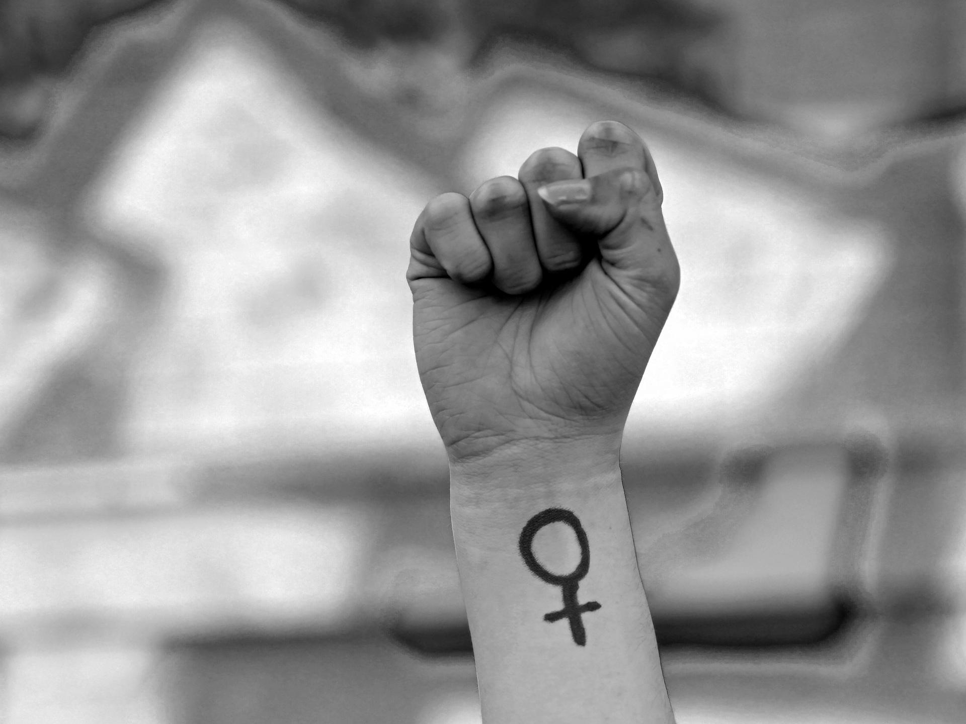 Eine Frau hebt ihre Faust in die Höhe. Auf ihrem Arm ist das Venussymbol zu sehen, das für das weibliche Geschlecht und Frauenrechte steht. Das Foto ist in Schwarz-Weiß gehalten. Eine Frau hebt ihre Faust in die Höhe. Auf ihrem Arm ist das Venussymbol zu sehen, das für das weibliche Geschlecht und Frauenrechte steht. Das Foto ist in Schwarz-Weiß gehalten.