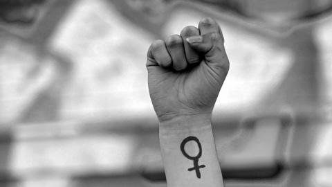Eine Frau hebt ihre Faust in die Höhe. Auf ihrem Arm ist das Venussymbol zu sehen, das für das weibliche Geschlecht und Frauenrechte steht. Das Foto ist in Schwarz-Weiß gehalten.