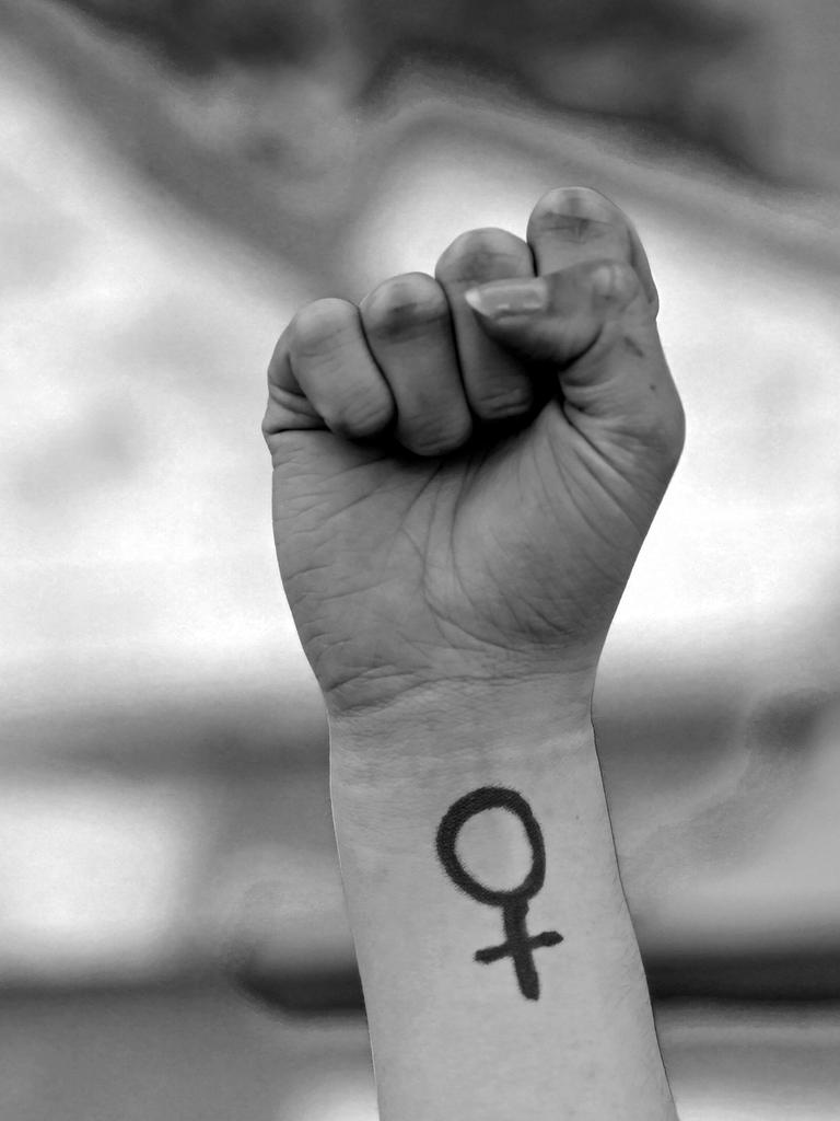 Eine Frau hebt ihre Faust in die Höhe. Auf ihrem Arm ist das Venussymbol zu sehen, das für das weibliche Geschlecht und Frauenrechte steht. Das Foto ist in Schwarz-Weiß gehalten. Eine Frau hebt ihre Faust in die Höhe. Auf ihrem Arm ist das Venussymbol zu sehen, das für das weibliche Geschlecht und Frauenrechte steht. Das Foto ist in Schwarz-Weiß gehalten.