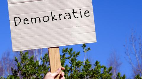 Pappschild mit der Aufschrift "Demokratie" wird hochgehalten, vor blauem Himmel.