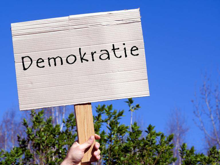 Pappschild mit der Aufschrift "Demokratie" wird hochgehalten, vor blauem Himmel.