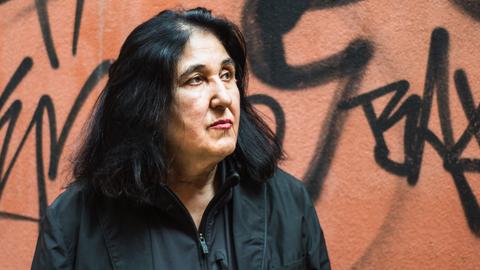 Porträt von Emine Sevgi Özdamar. Sie hat schwarze schulterlange Haare, trägt ein schwarzes Hemd und steht vor einer Wand mit Graffiti.
