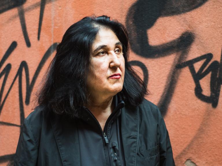 Porträt von Emine Sevgi Özdamar. Sie hat schwarze schulterlange Haare, trägt ein schwarzes Hemd und steht vor einer Wand mit Graffiti.