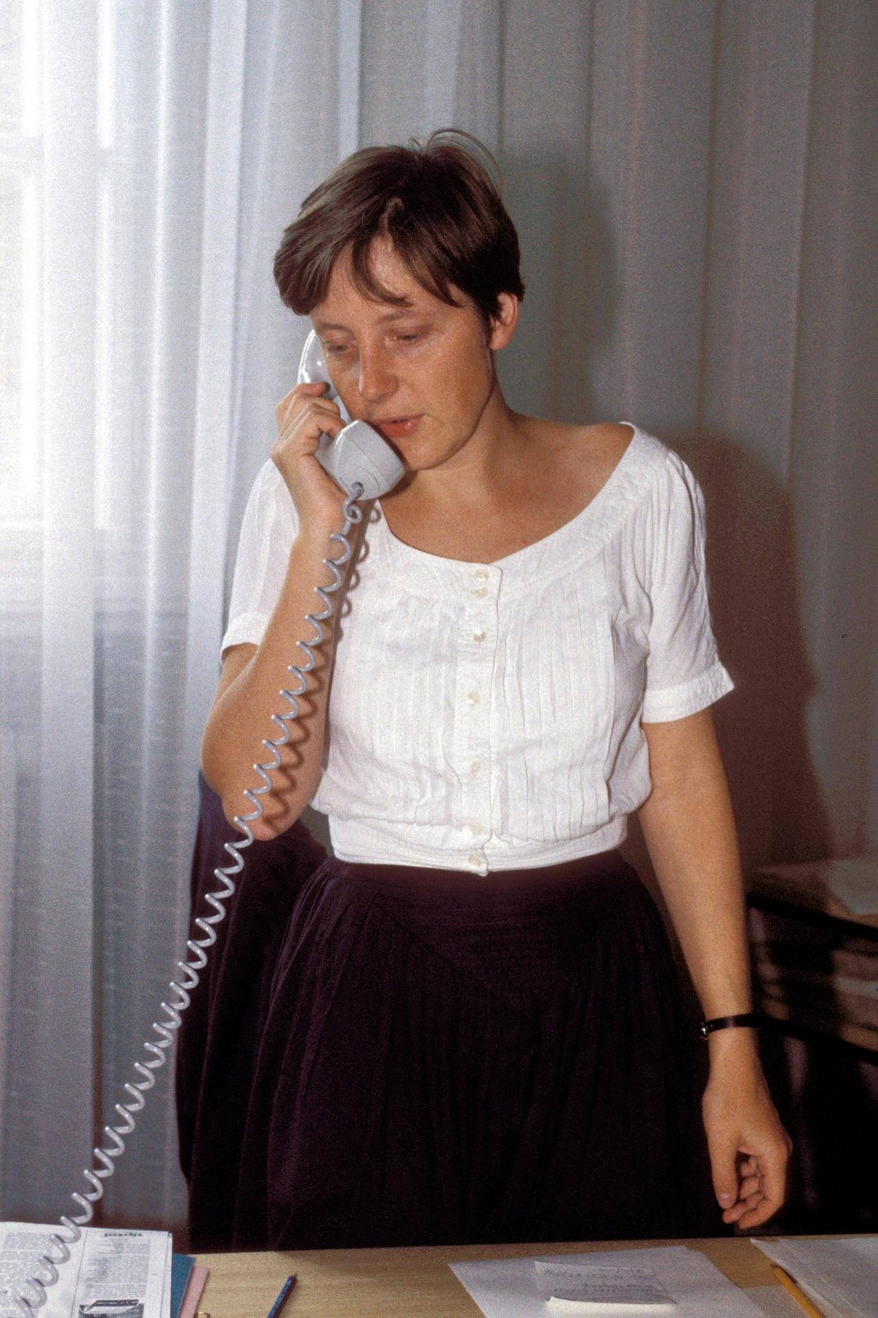 Eine junge Frau steht mit Telefonhörere in der Hand vor einem Schreibtisch
