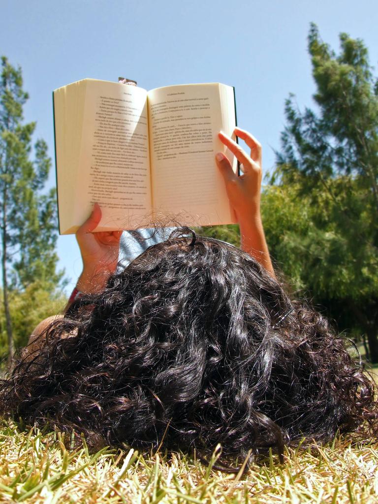 Junges Mädchen liegt auf dem Rücken auf einer Parkwiese und liest ein Buch.