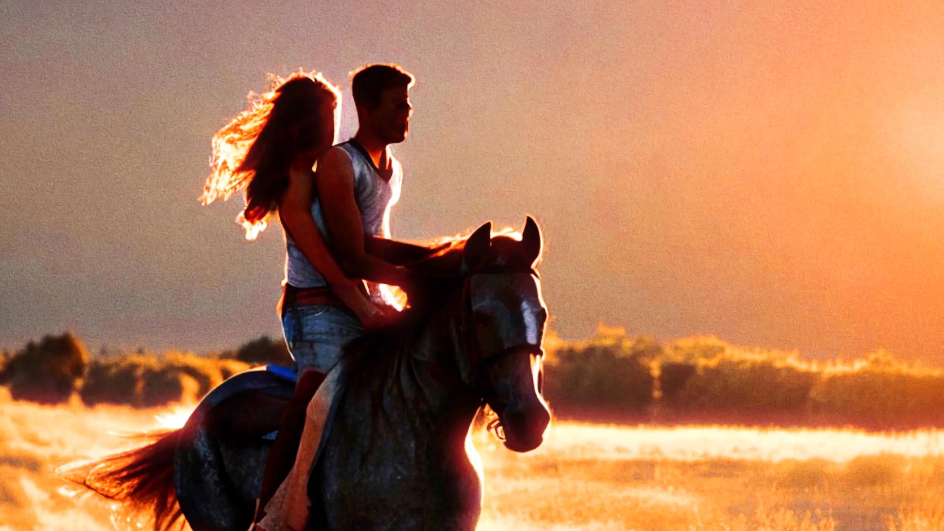 Eine junge Frau sitzt hinter einem jungen Mann auf einem Pferd. Sie reiten durch das Abendlicht. 