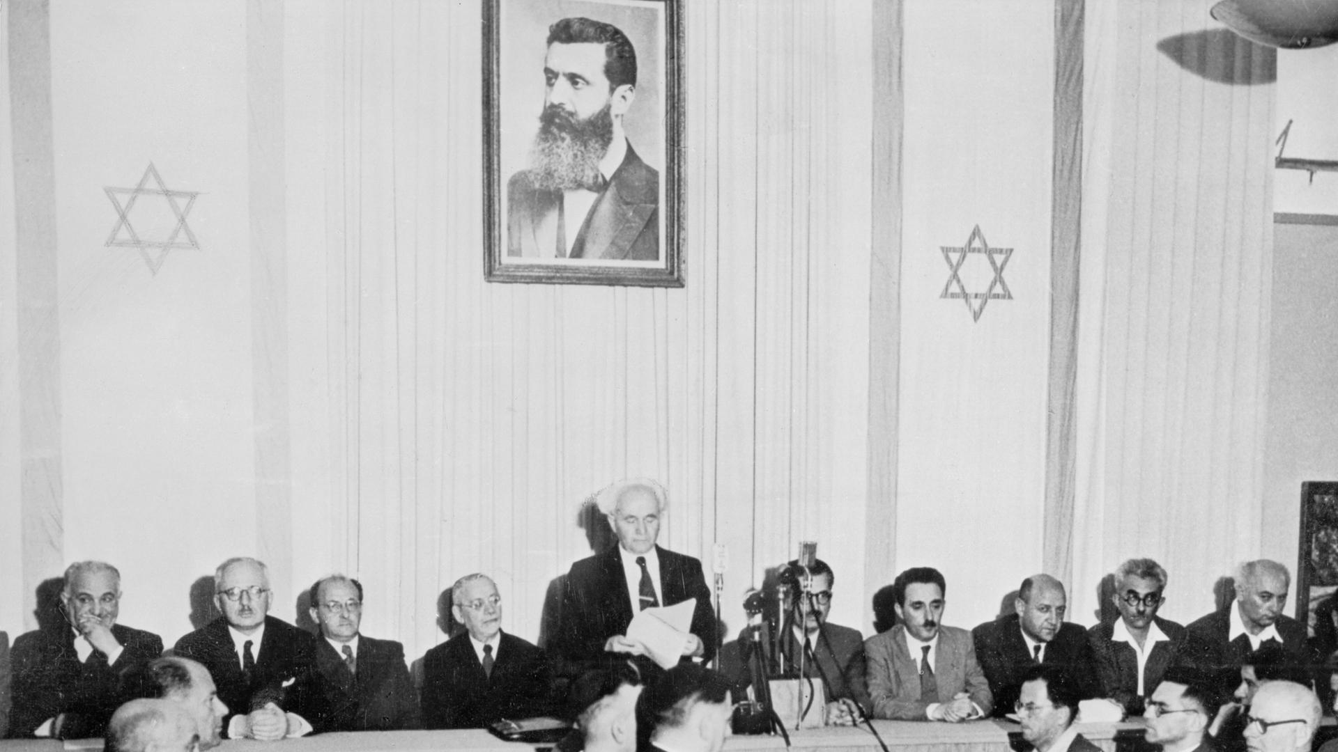 Israels erster Ministerpräsident Ben Gurion ruft die Gründung des Staates Israel aus. Er steht hinter einem Tisch. Zu seinen Seiten sitzen je vier Männer. Hinter ihm ein Bild von Theodor Herzl und zwei Israelflaggen.